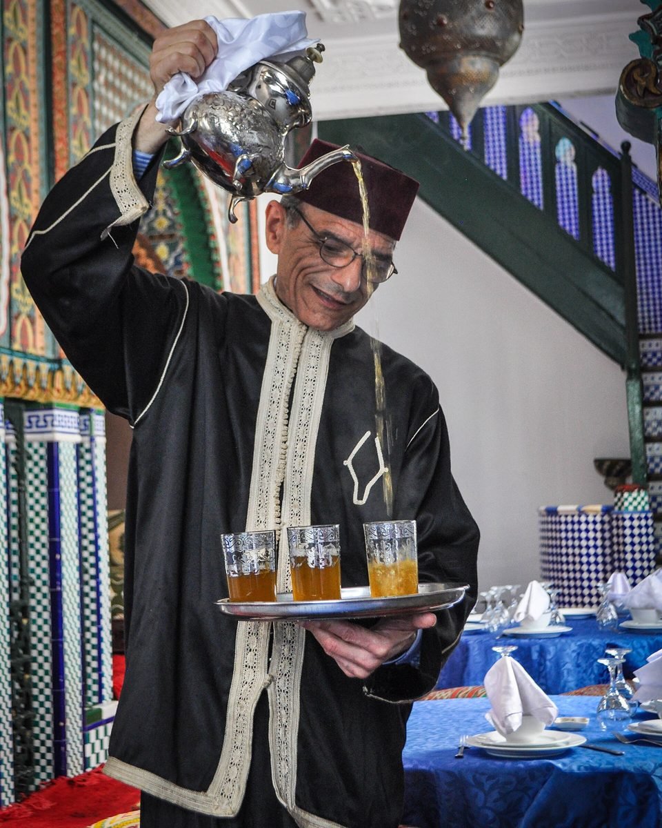 A waiter in a red fez and traditional jabador pours mint tea from a silver teapot into glasses on a tray