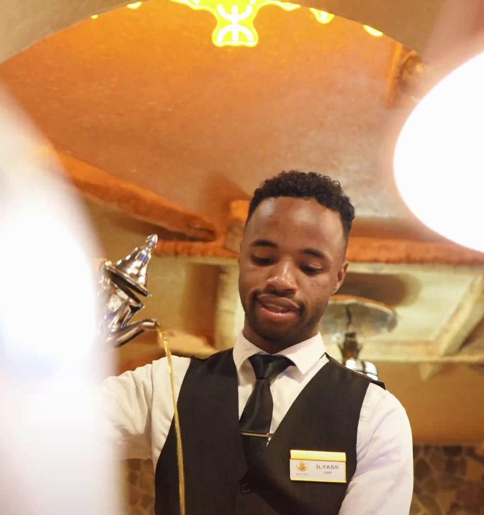 A smiling waiter in a black waistcoat and tie under the warm lamps of the dining room at La Table du Berbère
